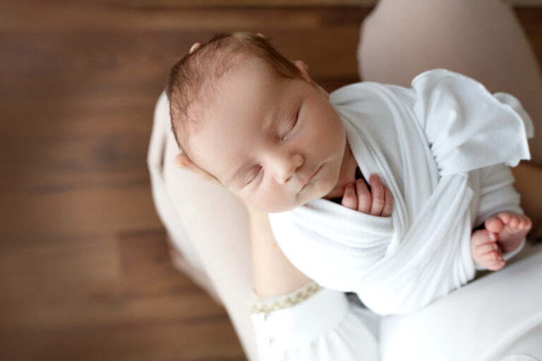 Top view of a newborn baby girl sleeping in a white baby wrap, lying on her mother's hands and knees. Against the background of dark brown natural wood. Portrait of a little girl 7 days, one week.